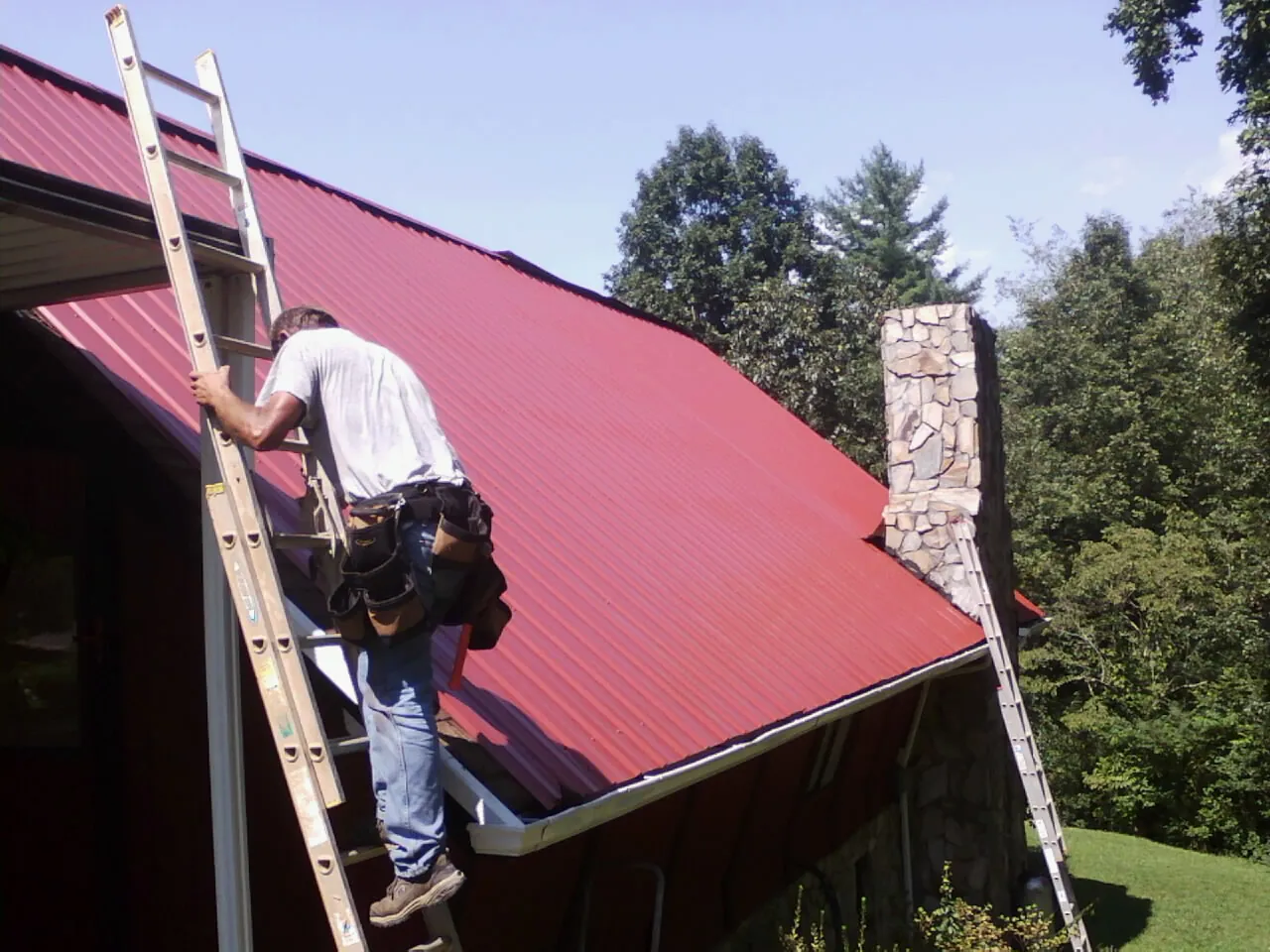Roofer on a ladder beside a red metal roof and stone chimney — Soffit & Fascia Repair by Roofing Brothers in Carter Lake
